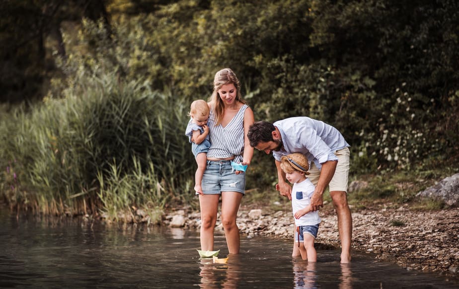 / canva – Young family with two toddler children outdoors by the river in summer, playing.